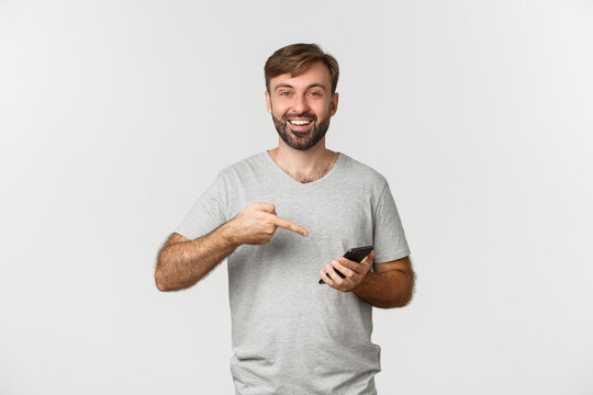 Portrait Of Excited Smiling Man In Gray T-shirt, Pointing At Mobile Phone, Showing Application, Standing Over White Background