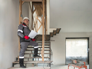 The Builder stands on the stairs and looks up. Construction engineer with drawings in his hands on the stairs. A man in a construction uniform and helmet in a renovated room.