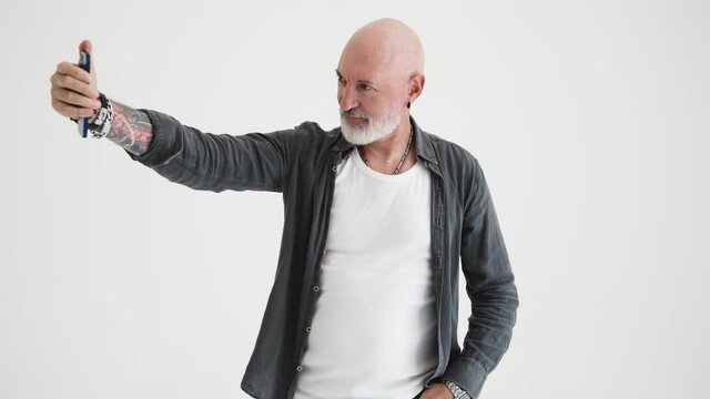 A handsome bald mature man takes selfie photo on his mobile standing isolated over white background in the studio