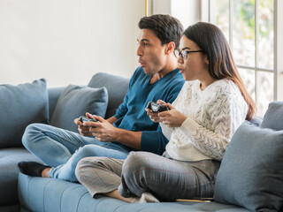 Handsome Caucasian man and beautiful Asian woman lovers sitting in house living room and using joystick to play game togehter with happiness and joyful. Diverse couple and stay at home concept