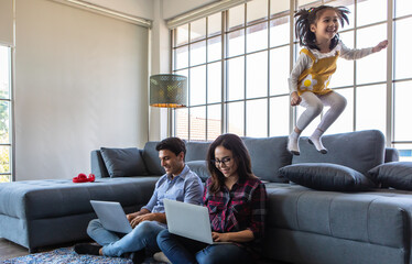 Mix race family lovers, Caucasian husband and Asian wife, parents working with laptop computers on floor while little naughty girl play and jump on sofa with fun and excitement. Work at home concept - 452019824