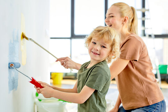 side view on positive family mother and child son making repairs, paint the wall at home. blonde caucasian lady teach kid boy to paint, friendly family in new apartment house. focus on child