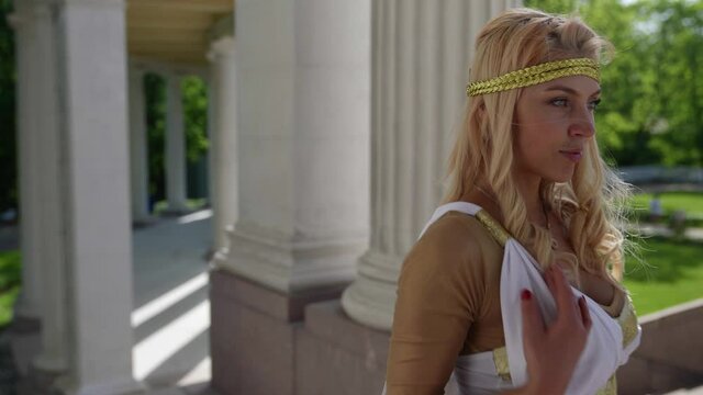 portrait of a blonde with big breasts and in a Greek chiton against the background of blurred park buildings