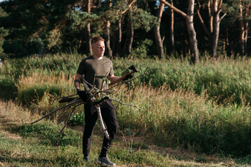 Photo of a Blond Man on a Summer Camping Activity with firewood for bonfire. Travel location of popular tourist outdoor. View of beautiful Forest Landscape. Vacation in the lake in a camp with a tent