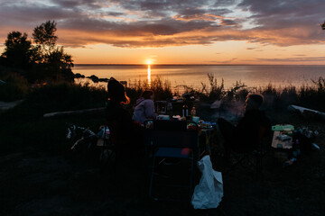 Photo of a Summer Camping Activity. Travel location of popular tourist outdoor. View of beautiful Sea and Forest Landscape. A group of friends on vacation by the lake in a camp with a bonfire and tent