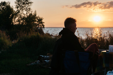 Photo of Man in a Summer Camping Activity. Travel location of popular tourist outdoor. View of beautiful Sea Landscape. Vacation by the lake in a camp with a bonfire and tent