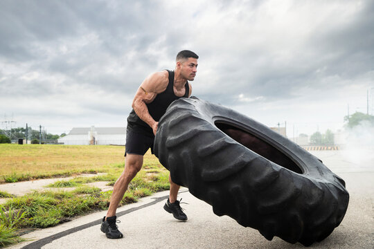 Muscular Hispanic man wearing tank top with Strong tattoo on his bicep, flipping tractor tire