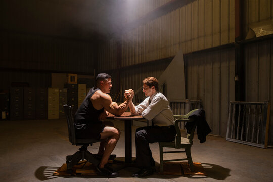 Two men preparing to have miss-matched arm wrestling contest in dingy warehouse 