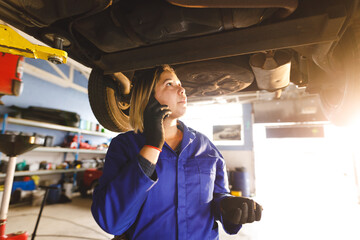 Mixed race female car mechanic wearing overalls, inspecting car, talking by smartphone