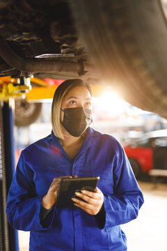 Mixed race female car mechanic wearing face mask and overalls, making notes on clipboard