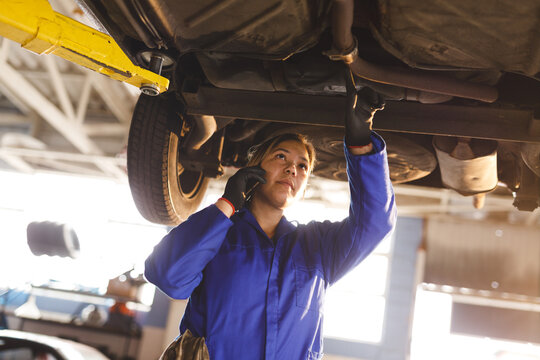 Mixed race female car mechanic wearing overalls, inspecting car, talking by smartphone
