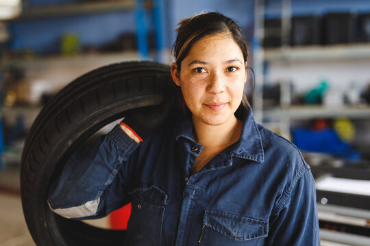 Mixed race female car mechanic wearing overalls, holding tire, looking at camera