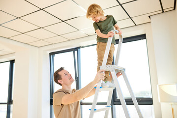 Home makeover, decoration. Little son helping father with building work, standing on ladder at...