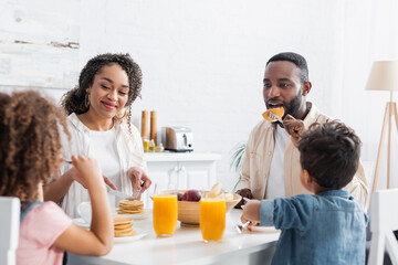 Obraz na płótnie Canvas african american couple having breakfast with blurred children in kitchen