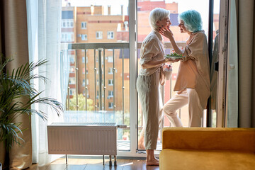 Side View Portrait Of Lovely Caucasian Lesbian Couple Enjoying Breakfast At Home Near Window, Relaxed And Happy In The Morning, Looking At Each Other, Smiling, In domestic Clothes, Women In love