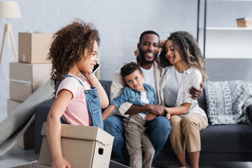 african american kid with cardboard box talking on smartphone near blurred family