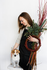 A beautiful Caucasian girl with loose hair and an apron is holding branches and a fir tree in her hands. There is a Jack Russell Terrier dog standing nearby. New Year and winter holidays.