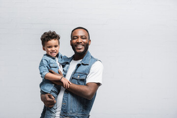 happy african american man holding kid while looking at camera on grey