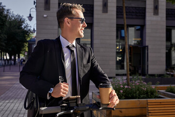 business and people concept. young businessman in sunglasses riding electric scooter outdoors, taking a break alone outdoors, drinking cup of coffee, enjoy leisure time at good day, feeling happy