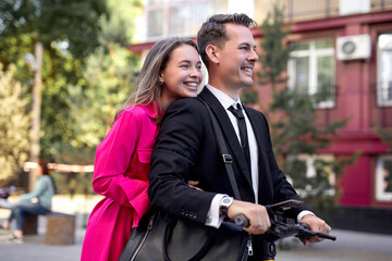 cheerful people, couple in elegant clothing driving electric scooter on city street, side view portrait of beautiful man and woman in suit and dress having fun, enjoying walk on scooter, have rest