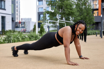 Fototapeta na wymiar Fitness lifestyle. Young smiling fat overweight african woman in black tracksuit doing push ups, side view sporty young lady at summer day in sport ground. Healthy life concept. Copy space