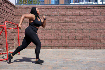 African fat woman jogging during cardio workout for burning calories outdoors in city at day time, side view, copy space. Young obese lady in black sportive tracksuit is running forward, focused. - obrazy, fototapety, plakaty