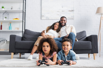 KYIV, UKRAINE - JANUARY 25, 2021: blurred african american couple smiling on sofa near kids gaming on floor