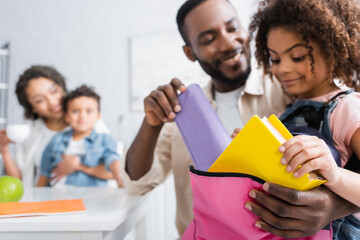 african american man helping daughter packing backpack near blurred family in kitchen