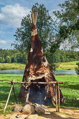 Indian wigwam covered with cow skins, bonfire and spit in the shade of trees © Alex
