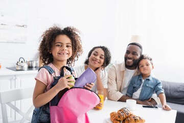 cheerful african american girl with pencil case and backpack near happy family in kitchen