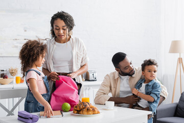 african american woman packing backpack of daughter in kitchen