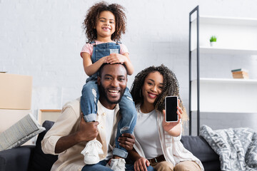 african american woman showing smartphone with blank screen near happy husband and daughter