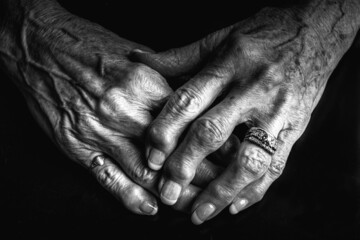 A close up black and white image of an elderly woman's arthritic hands. She is wearing a diamond ring and her knuckles are swollen. Her hands are gently placed on each other. Her nails are painted. 