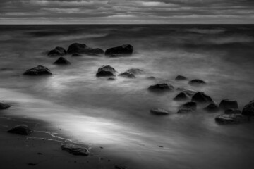 A moody black and white image of waves rolling into a rocky shore. A long exposure created a misty, dreamy feeling coupled with dark clouds. The image creates a feeling of peace and tranquility.  - Powered by Adobe
