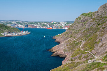 Fototapeta na wymiar A footpath, hiking trail or path along a hillside. The cliff is rocky with grass patches. The city of St. John's, Newfoundland, is in the background on a sunny day. The sky is bright blue. 