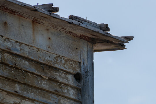 The exterior corner of an old wooden shed. The roof of the building is worn, rotten, and has grey textured boards on the wall with pieces of wood nailed to the roof. The eve extends over the building.