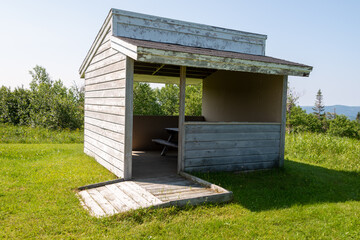 A wooden camp cookhouse with a picnic table inside the building at a campground. The wood board structure is in the middle of a green grass garden. The outdoor dining shelter area is empty. 