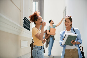 Female students read lecture and drink coffee in university hallway.