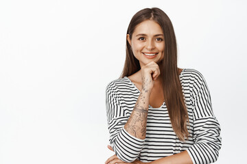 People portraits. Young woman smiling, looking with interest at camera, thinking of smth, hold hand on chin, standing against white background