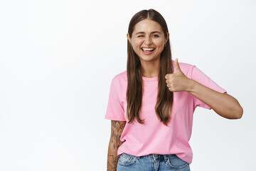 Obraz na płótnie Canvas Winking girl smiling and showing thumbs up, wearing pink tshirt, standing over white background