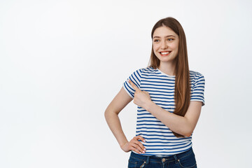 Obraz premium Happy and confident girl pointing, looking left with pleased smile, holding hand on waist, standing against white background
