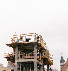 Building under construction with workers on top. Concrete and wooden framing. Structure against white sky background.