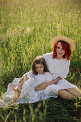 Fototapeta premium mother with red hair and a hat is sitting in a green field with grass with her daughter of four years sunset