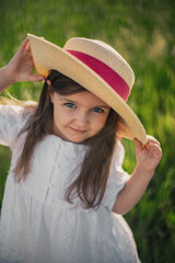 portrait of a beautiful girl in a straw hat standing in a field with green grass at sunset and looking the camera