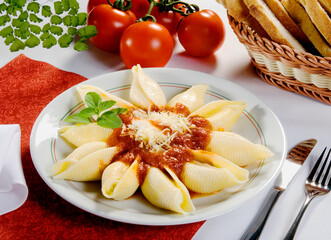 Conchiglione, massa típica da cozinha italiana, ao molho de tomate com queijo parmesão com cesta de pão italiano na mesa decorada.
