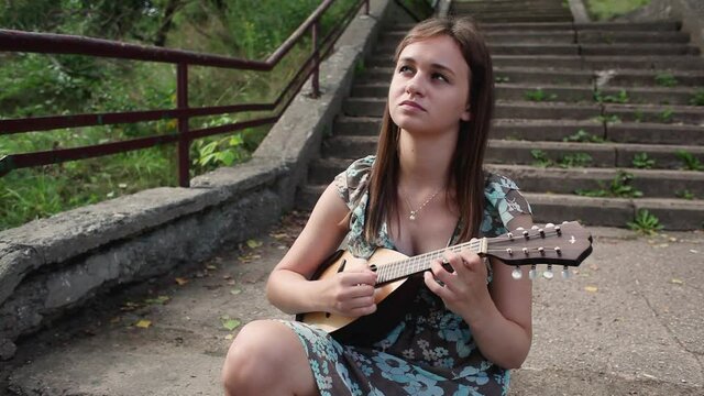 Young beautiful girl playing a musical instrument mandolin while sitting on the steps of the stairs . High quality FullHD footage