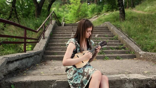 Young beautiful girl playing a musical instrument mandolin while sitting on the steps of the stairs . High quality FullHD footage