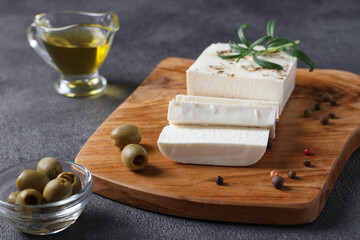 Feta cheese is located on a wooden board with olives, rosemary and olive oil on a dark background