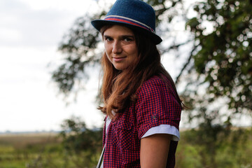 Defocus close up portrait of a smiling young woman with brown hair wearing a hat outdoors. Green nature background. Side view. Out of focus