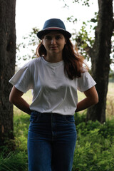 Defocus portrait of a sad young woman with brown hair wearing a hat outdoors. Green nature background. Looking at camera. Women standing at dark park. Vertical. Hands behind back. Out of focus